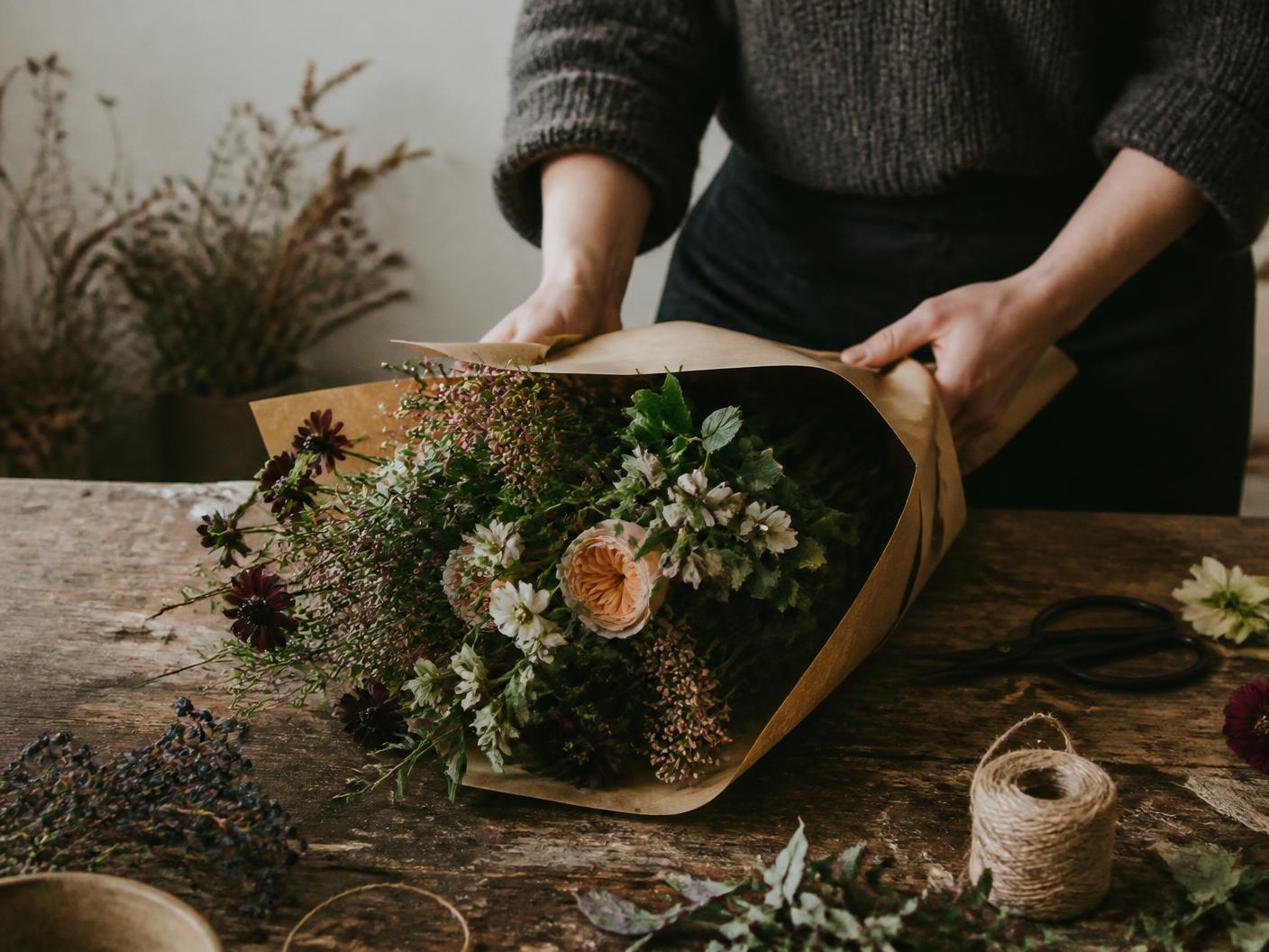 Hands wrapping a bunch of seasonal flowers in brown craft paper on a worn wooden table